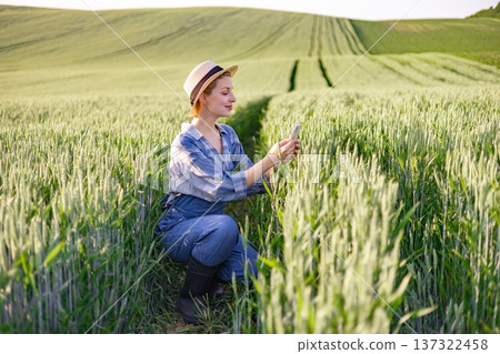 A woman in overalls and a hat crouches in a lush green wheat field, examining her smartphone A woman in overalls and a hat crouches in a lush green wheat field, examining her smartphone 137322458