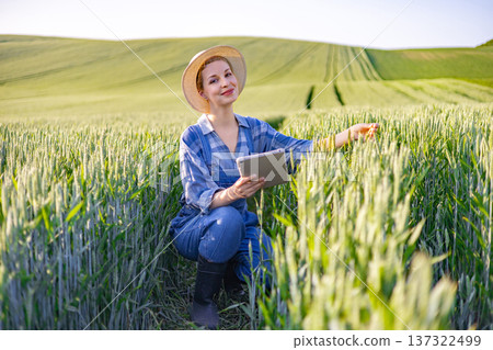 A woman in overalls and a straw hat crouches in a lush green wheat field, holding a tablet and examining the crops with a smile A woman in overalls and a straw hat crouches in a lush green wheat field, holding a tablet and examining the crops with a smile 137322499