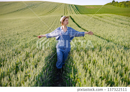 A young woman wearing overalls and a hat walks through a lush green wheat field, holding a few stalks of wheat in her hand A young woman wearing overalls and a hat walks through a lush green wheat field, holding a few stalks of wheat in her hand 137322511