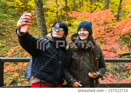 Two Russian women, aged 50 and 37, taking a selfie together in an autumn forest. Smiling friends enjoying nature, social media, and technology outdoors. Two Russian women, aged 50 and 37, taking a selfie together in an autumn forest. Smiling friends enjoying nature, social media, and technology outdoors. 137322710