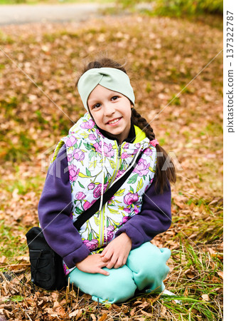 Russian girl sitting on the ground in autumn park, smiling softly in floral vest and green headband, surrounded by fallen leaves and warm seasonal tones. Russian girl sitting on the ground in autumn park, smiling softly in floral vest and green headband, surrounded by fallen leaves and warm seasonal tones. 137322787