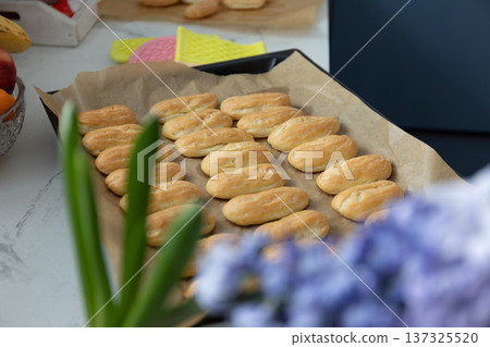 Freshly baked eclairs on parchment lined baking sheet in kitchen. Homemade custard filled French pastries with herb garnish. Baking success and sweet dessert concept for home cooking inspiration. 137325520
