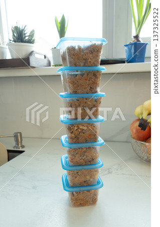 Tall stack of blue plastic containers filled with prepared pet food on white kitchen counter near window with plants and sink. Homemade meals for dogs and cats daily feeding routine. Pet nutrition Tall stack of blue plastic containers filled with prepared pet food on white kitchen counter near window with plants and sink. Homemade meals for dogs and cats daily feeding routine. Pet nutrition 137325522