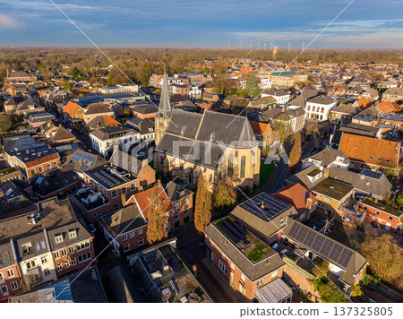 High angle aerial view of a historic European town featuring a prominent Gothic church with a tall spire, traditional brick houses with solar panels, and a scenic horizon under a soft sunset sky. High angle aerial view of a historic European town featuring a prominent Gothic church with a tall spire, traditional brick houses with solar panels, and a scenic horizon under a soft sunset sky. 137325805