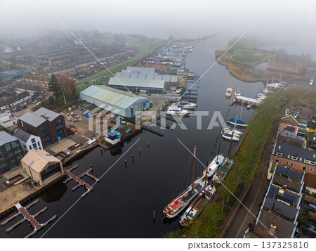 Aerial view of a misty morning at Muiden harbor, Netherlands. Features historic boats, modern shipyards and the medieval Muiderslot castle shrouded in fog Calm canal atmosphere with Dutch architecture Aerial view of a misty morning at Muiden harbor, Netherlands. Features historic boats, modern shipyards and the medieval Muiderslot castle shrouded in fog Calm canal atmosphere with Dutch architecture 137325810