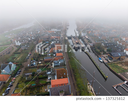Aerial drone view of a historic Dutch town with a long canal and traditional houses. Foggy morning over the water and rooftops of Muiden. Atmospheric urban landscape in the fog. Aerial drone view of a historic Dutch town with a long canal and traditional houses. Foggy morning over the water and rooftops of Muiden. Atmospheric urban landscape in the fog. 137325812