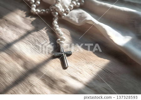 Pearl beads rest softly on fabric. Cross pendant glints under gentle sunlight. Warm light casts soft shadows across the scene. White cloth flows with natural folds 137328593