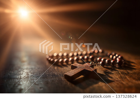 Warm sunlight shines behind wooden cross. Rosary beads rest softly on dark surface. Golden rays create dramatic spotlight effect. Cross and beads glow with spiritual warmth. Image evokes peace, faith Warm sunlight shines behind wooden cross. Rosary beads rest softly on dark surface. Golden rays create dramatic spotlight effect. Cross and beads glow with spiritual warmth. Image evokes peace, faith 137328624
