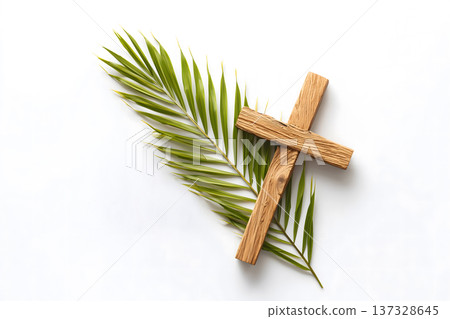 Simple wooden cross rests on green palm frond. Minimalist composition against plain white background. Natural textures highlight spiritual symbolism. Soft lighting enhances serene, peaceful mood 137328645