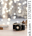 Black and white film camera aside books and vase on wooden table with blurred lights in background. 137328971