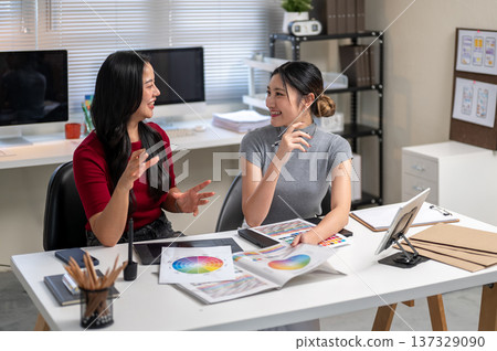 Asian woman holding color chart talking to coworker sitting with working material on table in office Asian woman holding color chart talking to coworker sitting with working material on table in office 137329090