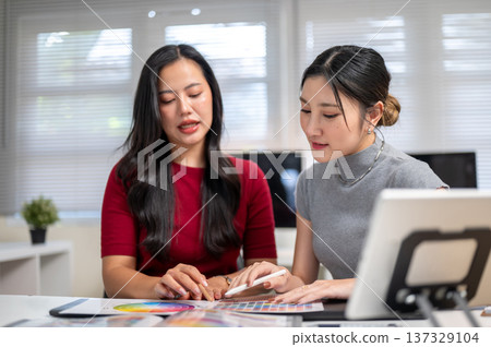 Asian woman explaining color chart to friend or coworker aside tablet on table in home office studio Asian woman explaining color chart to friend or coworker aside tablet on table in home office studio 137329104