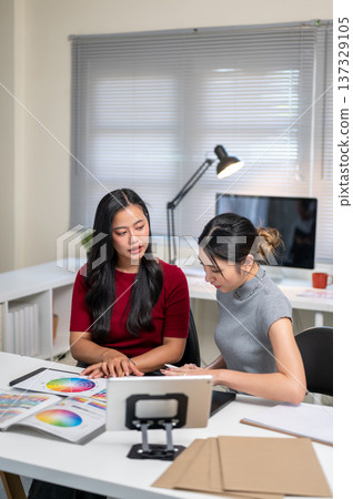 Asian woman talking to friend coworker over color chart aside tablet at table in home office studio. Asian woman talking to friend coworker over color chart aside tablet at table in home office studio. 137329105