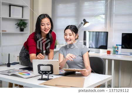 Pretty asian woman pointing at tablet sits on table aside smiling friend coworker in office studio. Pretty asian woman pointing at tablet sits on table aside smiling friend coworker in office studio. 137329112