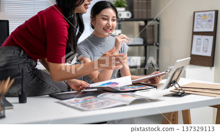 Pretty asian woman and friend or coworker talking while looking at tablet on table in office studio. 137329113