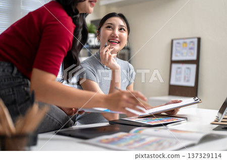 Pretty asian woman holding clipboard looking at a talking friend or coworker sitting on office table 137329114