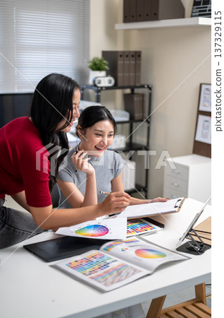 Pretty asian woman and a friend or coworker talking and looking at tablet on table in office studio. 137329115