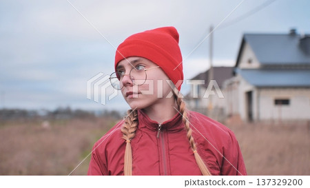 Portrait of a pensive teenage girl wearing a red beanie and glasses, standing in a field and looking away Portrait of a pensive teenage girl wearing a red beanie and glasses, standing in a field and looking away 137329200