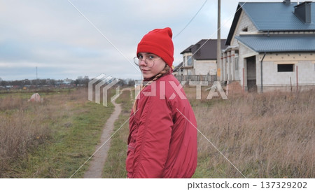 Young girl dressed in vibrant red clothes walking along a country road, surrounded by suburban houses amidst the beauty of autumn 137329202