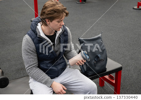 Young man sitting on gym bench holding protein shake bottle Young man sitting on gym bench holding protein shake bottle 137330440