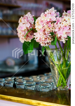 Fresh hydrangea flowers in clear vase with blurred cafe background. Floral decor, romantic interior and event decoration concept. Plastic flowers Fresh hydrangea flowers in clear vase with blurred cafe background. Floral decor, romantic interior and event decoration concept. Plastic flowers 137332599