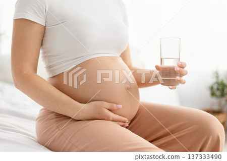 Pregnant woman sitting on bed holding belly and glass of water Pregnant woman sitting on bed holding belly and glass of water 137333490
