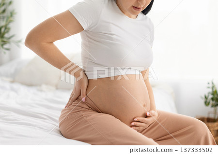 Pregnant woman holding belly and touching lower back while sitting on bed Pregnant woman holding belly and touching lower back while sitting on bed 137333502
