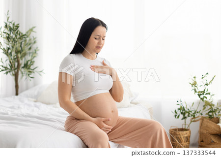 Pregnant woman sitting on bed touching chest and belly with discomfort Pregnant woman sitting on bed touching chest and belly with discomfort 137333544