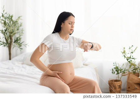 Pregnant woman sitting on bed checking smartwatch and holding belly at home. 137333549