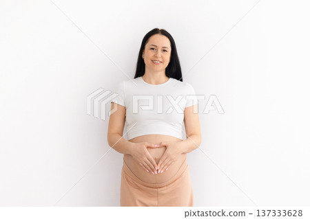 Pregnant young woman forming heart shape with hands on belly at home Pregnant young woman forming heart shape with hands on belly at home 137333628