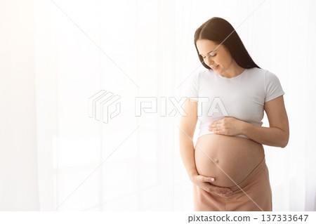 Young pregnant woman at home looking down at belly with gentle smile Young pregnant woman at home looking down at belly with gentle smile 137333647