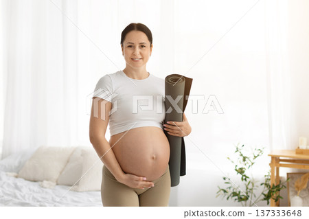 Smiling pregnant woman holding rolled yoga mat and touching belly at home Smiling pregnant woman holding rolled yoga mat and touching belly at home 137333648