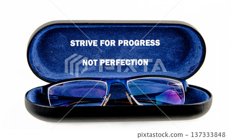 Blue spectacles rest peacefully in a velvet-lined case. Its open lid displays a timeless message: Pursue progress, not perfection on a bright white setting 137333848