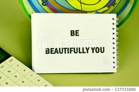 A pristine white notebook displays the powerful words BE BEAUTIFULLY YOU on a vibrant green desktop a partial calculator and colorful disk nearby A pristine white notebook displays the powerful words BE BEAUTIFULLY YOU on a vibrant green desktop a partial calculator and colorful disk nearby 137333880