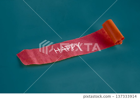 Crimson fabric ribbon unrolls on a rich teal background, revealing the word HAPPY in clear white letters, radiating cheer and positivity 137333914