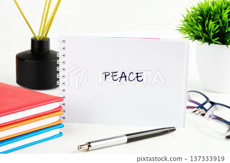 A white notebook displays the word Peace on a bright desk. Colorful books, a pen, diffuser, reading glasses, and a green plant complete the serene office setup A white notebook displays the word Peace on a bright desk. Colorful books, a pen, diffuser, reading glasses, and a green plant complete the serene office setup 137333919