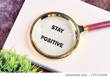 A magnifying glass rests on a pristine white notebook, clearly showing the phrase Stay Positive in bold letters, with a green plant nearby A magnifying glass rests on a pristine white notebook, clearly showing the phrase Stay Positive in bold letters, with a green plant nearby 137333936