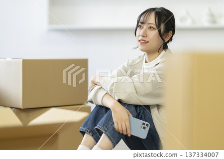 A young woman holding a smartphone sitting surrounded by cardboard boxes. Moving image. 137334007