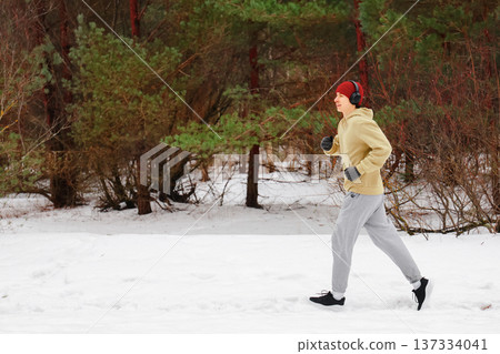 Side view of a young man jogging in a snowy forest. Male runner in headphones and warm sportswear listening to music during a winter workout outdoors. High quality photo Side view of a young man jogging in a snowy forest. Male runner in headphones and warm sportswear listening to music during a winter workout outdoors. High quality photo 137334041