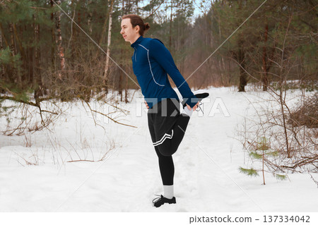 Young male runner stretching leg muscles in a snowy forest. Man warming up before jogging outdoors. Winter fitness and healthy lifestyle concept in cold weather. High quality photo Young male runner stretching leg muscles in a snowy forest. Man warming up before jogging outdoors. Winter fitness and healthy lifestyle concept in cold weather. High quality photo 137334042
