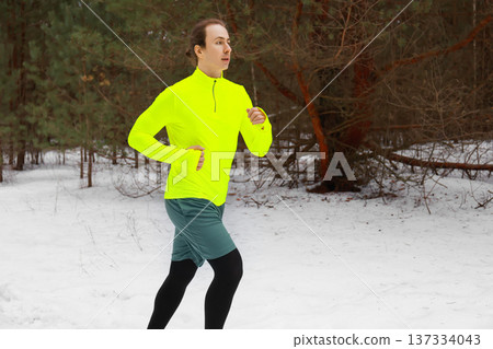 Side view of a male runner in neon sportswear jogging in a winter forest. Young man exercising outdoors on a snowy trail. Healthy lifestyle and cold weather fitness concept. High quality photo Side view of a male runner in neon sportswear jogging in a winter forest. Young man exercising outdoors on a snowy trail. Healthy lifestyle and cold weather fitness concept. High quality photo 137334043