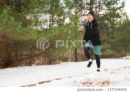 Young man jogging on a snowy trail in a pine forest. Winter outdoor fitness workout. Male runner exercising in cold weather wearing a black hoodie and beanie. High quality photo 137334045