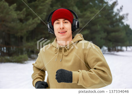 Happy young man jogging in a snowy winter forest wearing wireless headphones and a red beanie. Portrait of a male runner listening to music during an outdoor cold weather workout. High quality photo Happy young man jogging in a snowy winter forest wearing wireless headphones and a red beanie. Portrait of a male runner listening to music during an outdoor cold weather workout. High quality photo 137334046