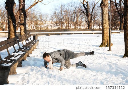 Young man slipping and falling on snow in a winter park. Clumsy guy stumbling outdoors, illustrating the risk of accidental injury and trauma in cold weather. High quality photo 137334112