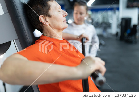 Senior woman exercising on gym machine with trainer Senior woman exercising on gym machine with trainer 137334129