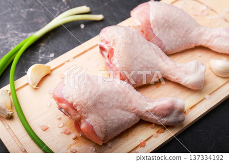 Raw chicken drumsticks arranged on a wooden board with green onions, garlic, and sea salt. Fresh poultry meat ingredients on a dark background ready for cooking. High quality photo 137334192