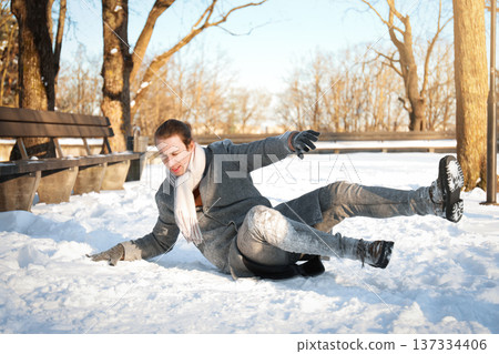 Young man slipping on ice and falling down in a snowy park. Winter accident, injury, and health insurance concept. Dangerous slippery road during a cold day. High quality photo 137334406