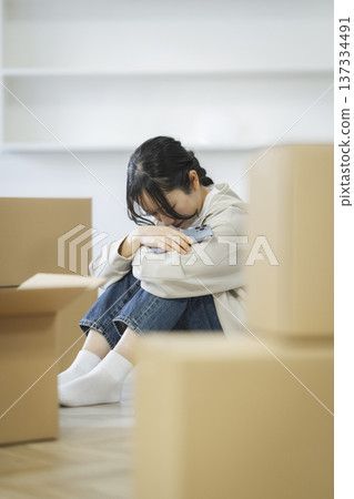 A young woman sits surrounded by moving luggage, holding her knees and holding a smartphone 137334491