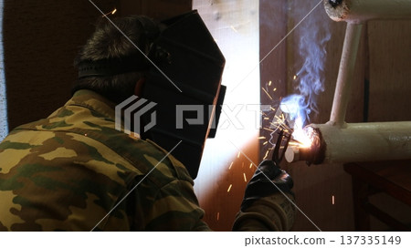 A welder, seen from behind, in uniform and a protective helmet, welds a section of a home heating system made of white-painted iron pipes. A man wearing a special mask welds a heating radiator. 137335149