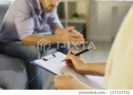 Therapist writing on clipboard as patient listens during private consultation 137335792